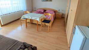 Une petite pièce propre et bien rangée, avec un lit, une table entourée de chaises, un radiateur et une fenêtre avec des rideaux.