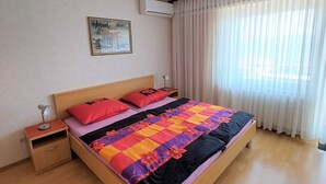 A neatly arranged hotel room with a wooden bed, a nightstand with a lamp, a wall-mounted air conditioner, and a window with sheer curtains.