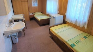 Une chambre de taille modeste, comprenant un lit simple, un petit bureau avec une chaise, un lavabo et un réfrigérateur.