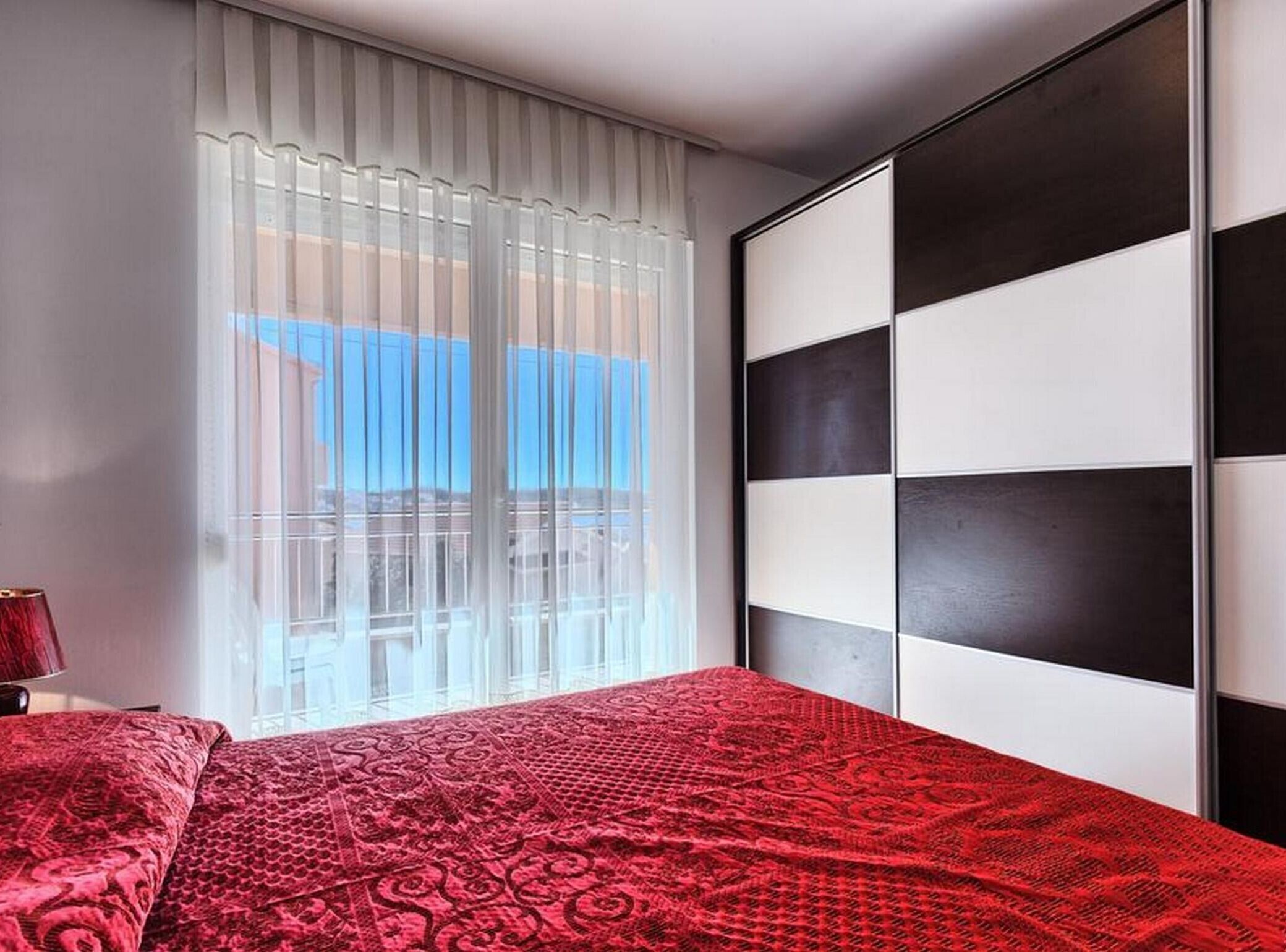 A bedroom with a large window, a bed with a red patterned bedspread, and a black and white headboard.