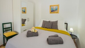 Una cama bien hecha con una colcha amarilla, dos almohadas y dos toallas enrolladas. Hay una silla verde con un cojín amarillo, una mesita con una lámpara y un cuadro en la pared.