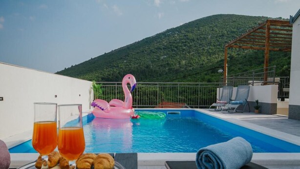 Escena junto a la piscina con un flotador de flamenco, una bandeja de cruasanes y vasos de jugo, y una toalla en una silla de descanso.