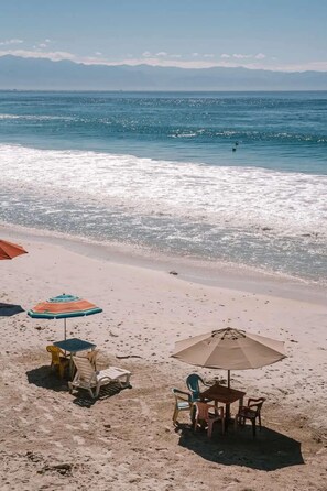 Ein Strand mit Tisch und Stühlen unter einem großen Sonnenschirm, und ein weiterer kleinerer Sonnenschirm in der Nähe.