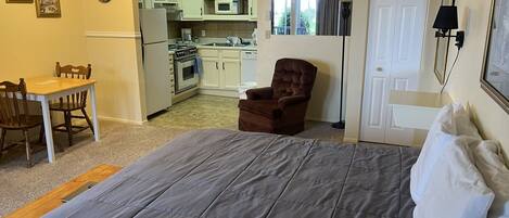 Une chambre avec un grand lit, une chaise, un bureau et, au fond, un coin cuisine.