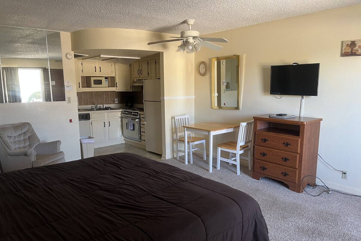 Ein kleines Zimmer mit Bett, Schreibtisch, Stuhl, Kommode, Fernseher und einer Küchenzeile.