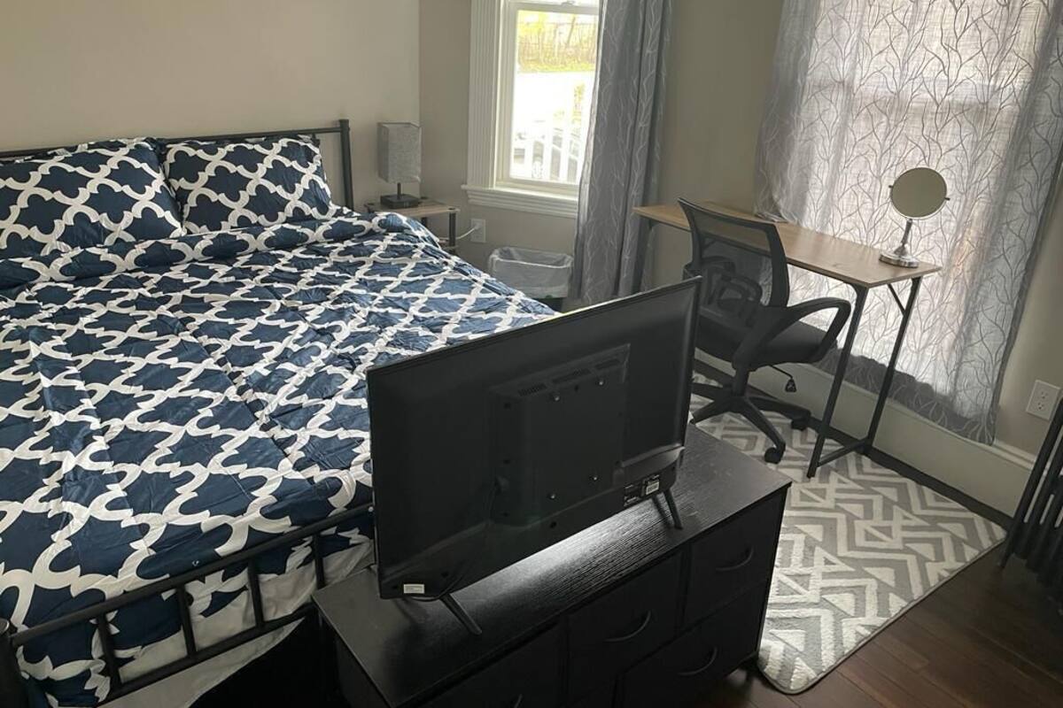 A bedroom with a bed, a desk, a chair, a TV, and a window with curtains.