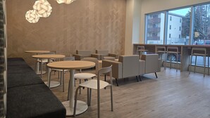 Una cafetería moderna con mesas y sillas redondas, un área de sofás y ventanas amplias con vista a la calle.