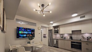 Una cocina moderna equipada con microondas, refrigerador y lavavajillas. Hay una zona de comedor con una mesa y sillas, y un televisor de pantalla plana en la pared que muestra varios canales.