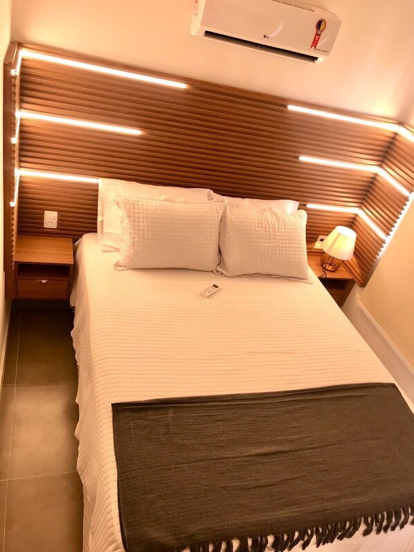 A neatly made bed with a white quilt and two pillows, a bedside lamp, and a wall-mounted air conditioner. - Apartamento Porto de Galinhas, Centro, Vista Mar, Ipojuca (Maracaipe Ipojuca)