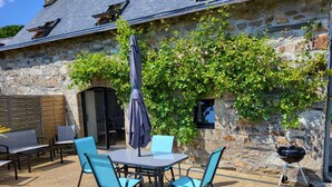 A stone building with a patio area featuring a table and chairs, a grill, and a covered umbrella. - Gite de Pennenez  (Plomodiern)