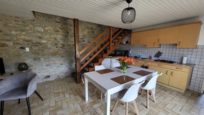 A dining area with a table, chairs, and a vase of flowers. There is a kitchen area with cabinets and a stove. A wooden staircase is visible. - Gite de Pennenez  (Plomodiern)