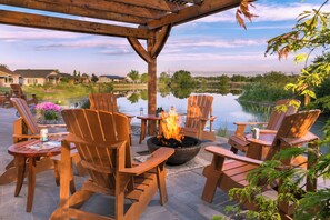 A patio with wooden chairs and tables, a fire pit, and a view of a lake and houses in the background. - Modern one bedroom in quiet lakeside retreat near wineries, dock, boats and more! (Zillah)