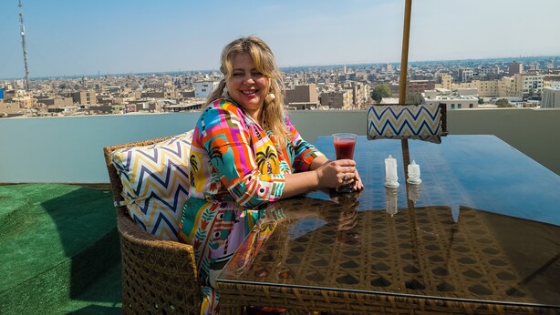 Una persona sentada en una mesa con vistas a la ciudad, un cojín estampado y una bebida.