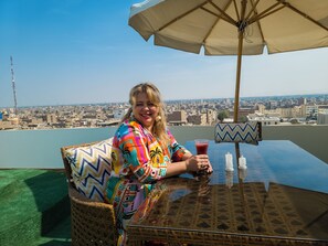 A person seated at a table with a city view, a patterned cushion, and a drink.