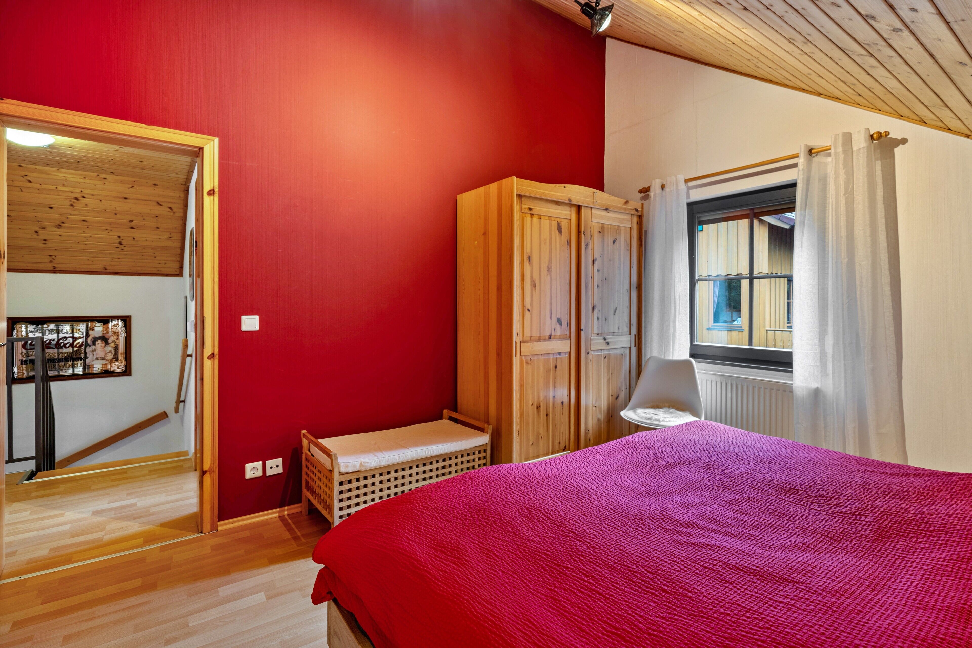 Une chambre avec une armoire en bois, un lit recouvert d’une couverture rouge, une fenêtre avec des rideaux et un plancher en bois.
