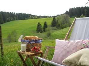 Uma mesa de madeira com pão em cesta, um copo de leite e uma tigela de morangos, com vista para um campo verde e uma floresta.