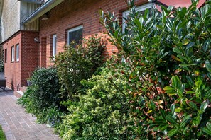 A red brick building with a brick pathway, dense green foliage, and a metal fence.