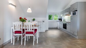 A modern kitchen with white cabinetry, a dining area with white chairs and red cushions, and a large window providing natural light. - Apartment 'Ferienwohnung Voglkoje' with Private Terrace, Shared Garden and Wi-Fi (Pellworm)