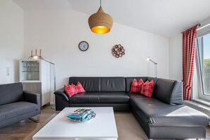 Uma sala de estar moderna com um sofá de couro preto, uma mesa de centro branca, um lustre tecido pendente, um relógio de parede e um objeto decorativo na parede.