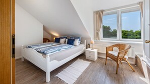 A bedroom with a bed, a nightstand, a wicker chair, a window with curtains, and a radiator. - Apartment 'Ferienwohnung Voglkoje' with Private Terrace, Shared Garden and Wi-Fi (Pellworm)