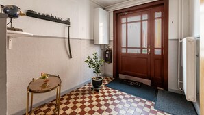 Un pasillo con piso a cuadros, una puerta de madera, un radiador, una mesa pequeña con una planta y una estantería con un tren modelo.