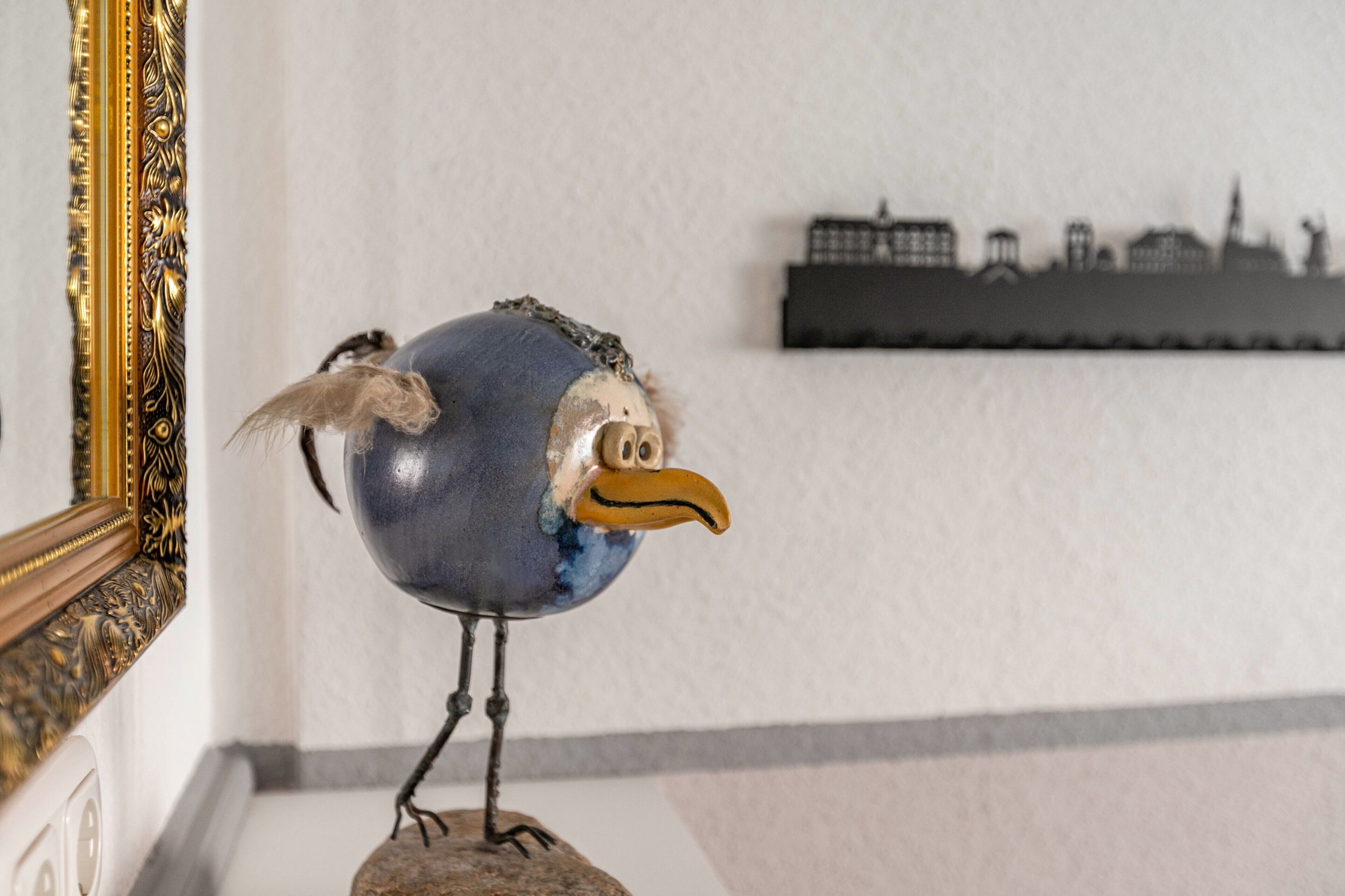 A bird sculpture on a pedestal in a room with a white wall and a framed picture.