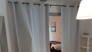 Una habitación con suelo de madera, cortinas blancas, una cama, un cuadro colgado en la pared y un estante blanco.