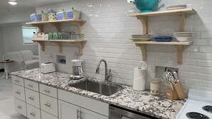 Une cuisine moderne dotée de plans de travail en granit, de meubles blancs et d’appareils électroménagers en acier inoxydable. Des étagères en bois, situées au-dessus de l’évier, servent à ranger la vaisselle et les verres.