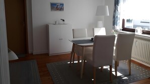 A room with a dining table, chairs, a cabinet, a lamp, and a window with curtains. - Apartment 'Ferienwohnung Edel' with Private Terrace and Wi-Fi (Wesselburen)