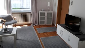 A modern room with a TV, a chair, a cabinet, a radiator, a window with curtains, and a small table. - Apartment 'Ferienwohnung Edel' with Private Terrace and Wi-Fi (Wesselburen)