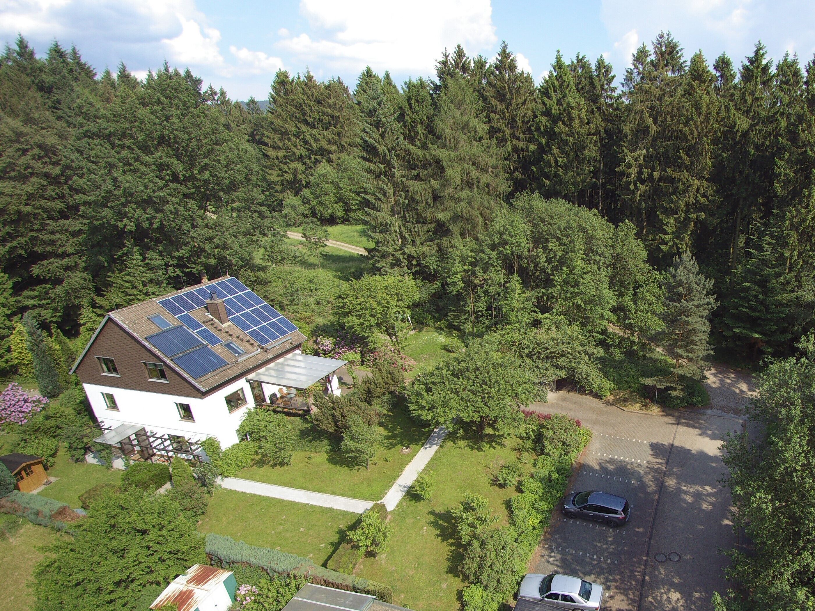 Une maison dotée d’un toit recouvert de panneaux solaires, entourée d’une forêt dense, avec une allée et des voitures garées.