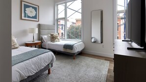A bedroom with two beds, a large window, a television, and a framed picture on the wall. - Bright Cozy Loft | Walk to PATH Shops + Dining (Hoboken)