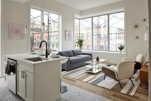 A modern kitchen with a built-in island, a gray sofa, and a glass coffee table. - Bright Cozy Loft | Walk to PATH Shops + Dining (Hoboken)
