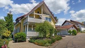 Ett tvåvåningshus med en balkong, omgivet av en trädgård med olika växter och blommor.