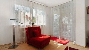 Ett modernt rum med en röd soffa, en golvlampa, tunna gardiner och en utsikt genom stora fönster.