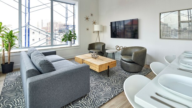 A modern living room with a grey sofa, a wooden coffee table, and a zebra-patterned rug. There are two potted plants, a floor lamp, and a dining area with white chairs and a table. - Bright Designer Loft  –  steps from PATH to NYC (Hoboken)