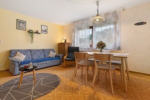 Een woonkamer met een bank, een eettafel, stoelen, een bijzettafeltje en een raam met gordijnen.
