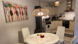 Un coin repas avec une table ronde, des chaises blanches, un tableau et une cuisine en arrière-plan.