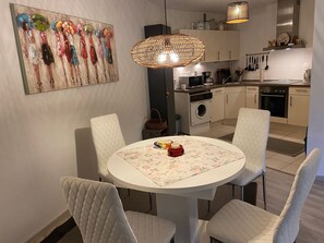 Un coin repas avec une table ronde, des chaises blanches, un tableau et une cuisine en arrière-plan.