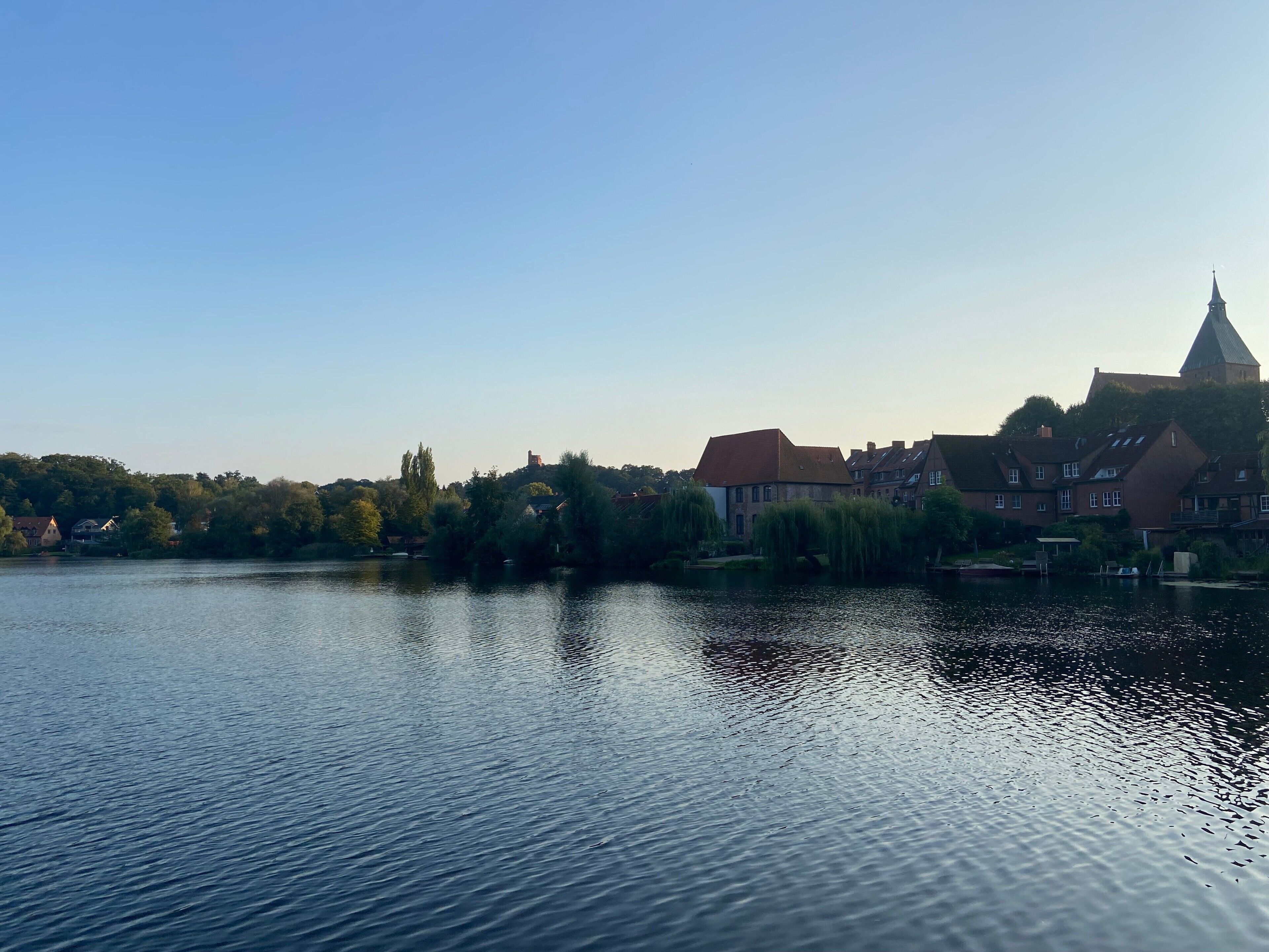 Ein ruhiger Fluss mit Gebäuden am rechten Ufer, darunter eine Kirche mit einem Turm.