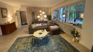 Un salon comprenant un canapé, un pouf et une table basse, une cheminée et une grande fenêtre avec des stores.