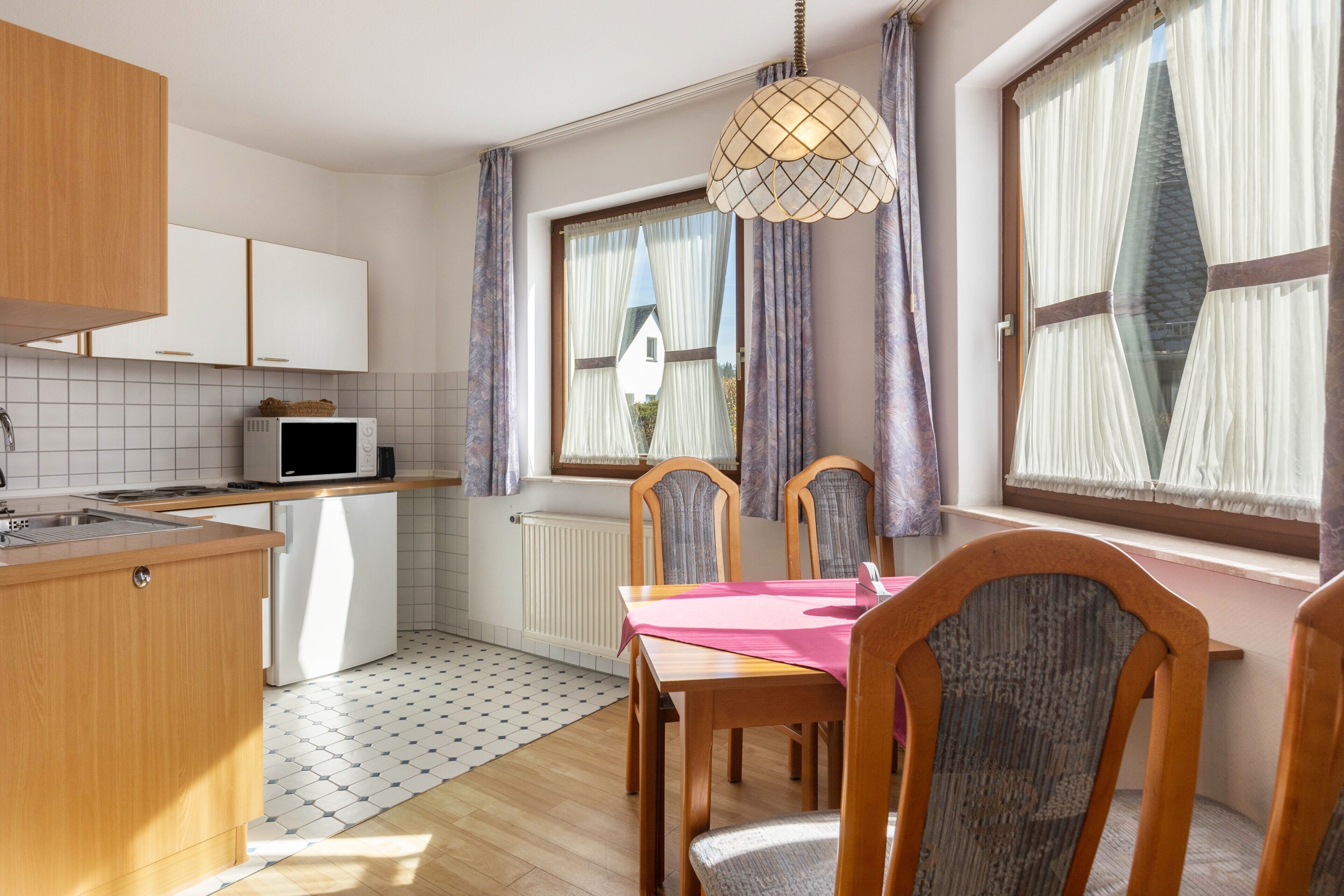 A compact kitchen with a microwave, wooden cabinets, a dining table with chairs, and a window with curtains.
