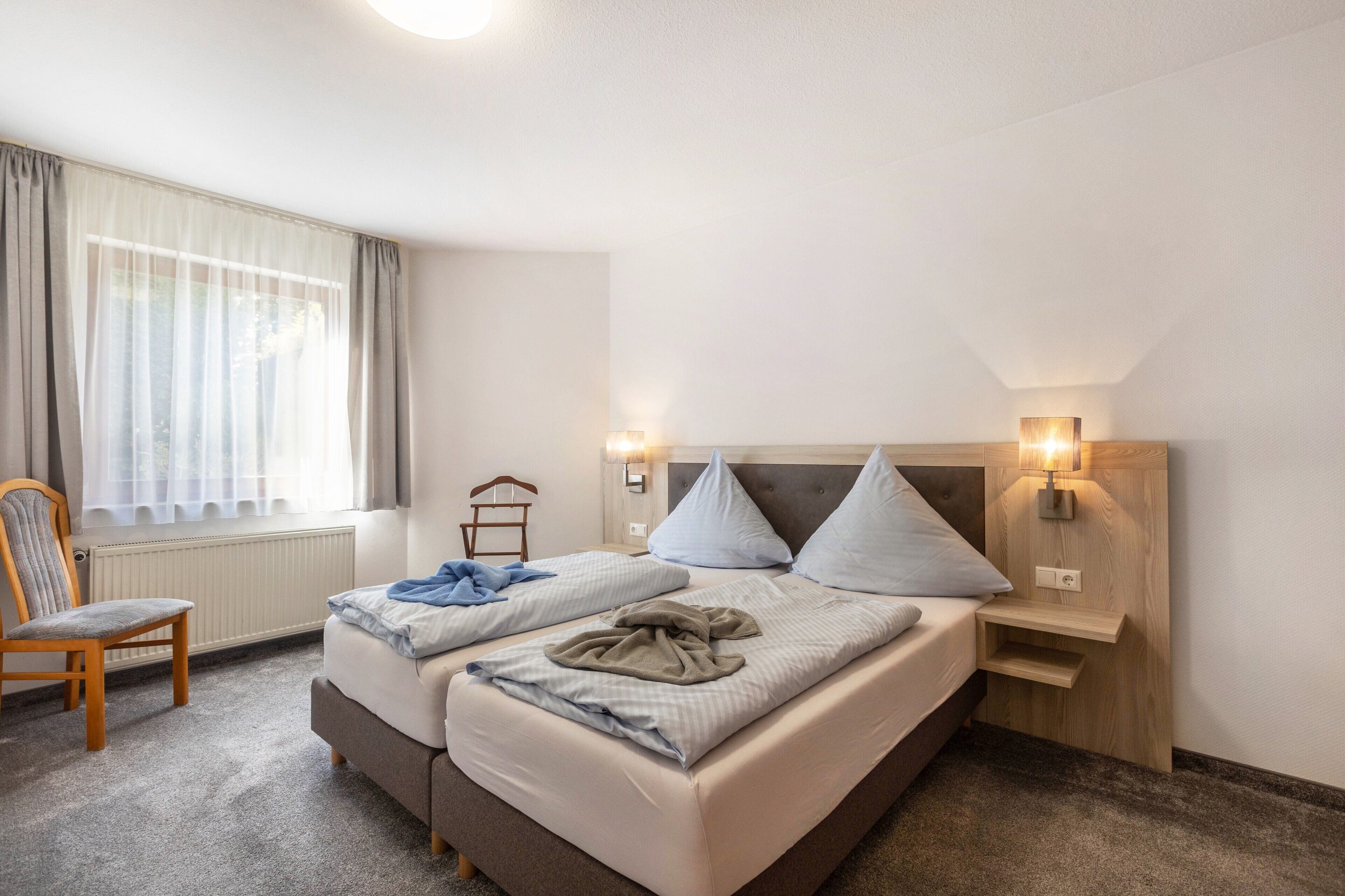 A hotel room with two beds, a chair, a nightstand with a lamp, and a window with curtains.