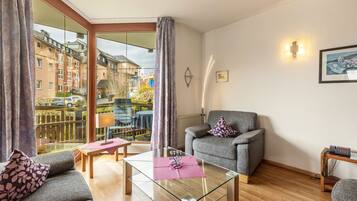 Ein modernes Wohnzimmer mit Glastisch, grauem Sofa und Sessel, ein Balkon mit Blick auf Gebäude und ein kleiner Tisch mit Lampe.