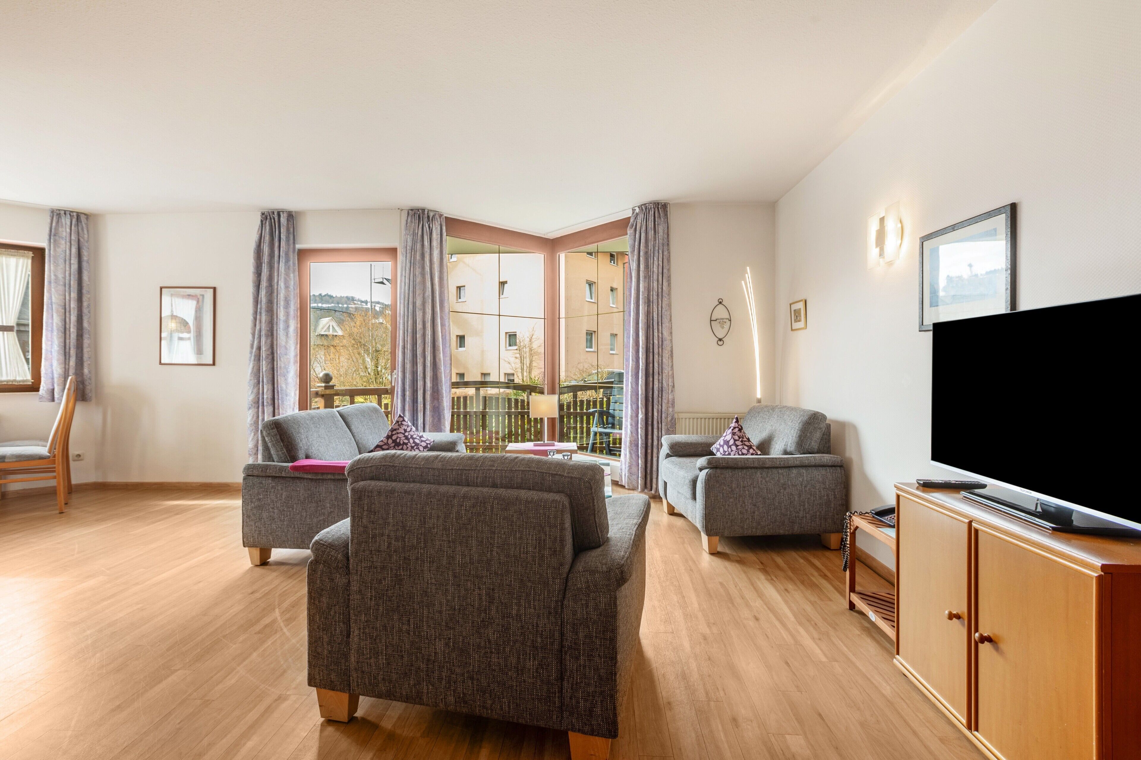 Ein Wohnzimmer mit zwei grauen Sofas, einem Holzkasten, einem Fernseher und einem Balkon mit Blick auf Gebäude.