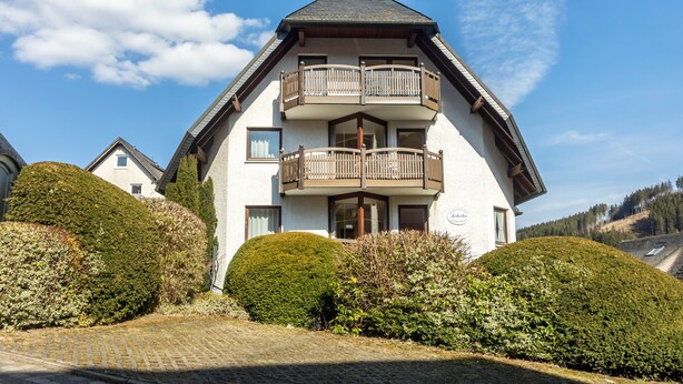 Un bâtiment à deux étages doté d’un balcon à l’étage supérieur, entouré de haies bien entretenues.