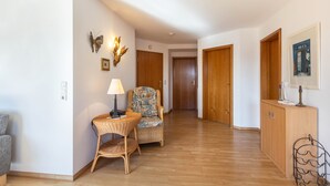 Un couloir avec un fauteuil en osier, une petite table avec une lampe, des portes en bois et un meuble sur lequel est posée une image.