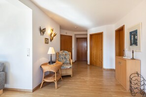 A hallway with a wicker chair, a small table with a lamp, wooden doors, and a cabinet with a picture.
