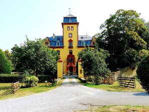 A large, yellow building with a central tower, a gravel driveway, and surrounding greenery.