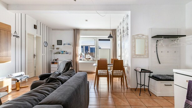 A modern living room with a sofa, chairs, a dining table, and a kitchen area with a refrigerator and hanging clothes rack. - Holiday Apartment 'Friedrichskoog Spitze' with Private Terrace and Wi-Fi (Friedrichskoog)